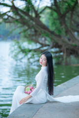 A young Vietnamese woman in a white ao dai standing by Hoan Kiem Lake, holding a bouquet of pink lotus flowers