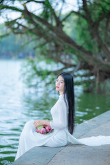 A young Vietnamese woman in a white ao dai standing by Hoan Kiem Lake, holding a bouquet of pink lotus flowers