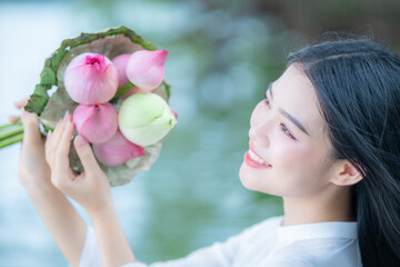 A young Vietnamese woman in a white ao dai standing by Hoan Kiem Lake, holding a bouquet of pink lotus flowers