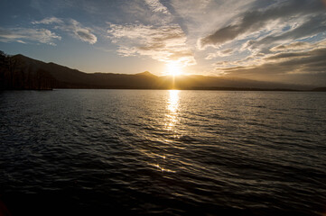 風に吹かれた湖の日暮れの場面。さざ波の水面に太陽光が反射し空にはドラマチックな雲の模様。