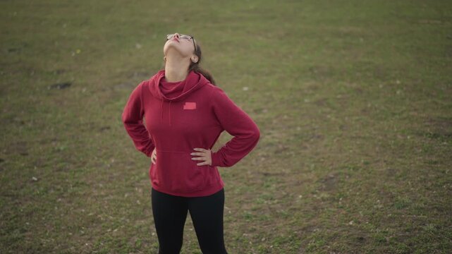 White Woman Performing Neck Mobility Stretches On Grass, Hands On Hips, Steady Breathing And Upward Rotations, Red Hoodie And Glasses, Focused Posture Work And Cool Open Field Setting