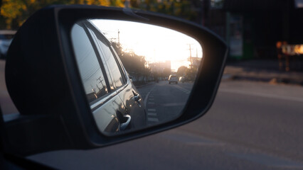 Side mirror view of black car. Close up on side car reflection in the mirror. With blurred of car driving on clear road with sunlight on the town faraway . Environment of evening time.