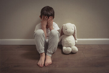 A lonely child sits on the floor hiding their face beside a soft toy, symbolizing sadness, fear, and emotional trauma from domestic abuse.