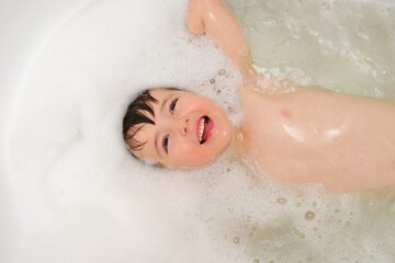 Fototapeta premium Young happy boy takes a bubble bath with white foam, looking up at camera. Concept of child hygiene and joyful bathing experience. Child aged 4 years (four years old)