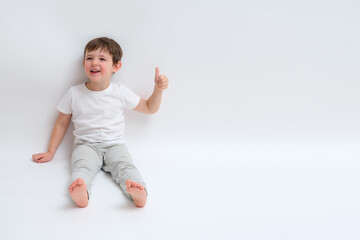 Smiling boy in a white t-shirt and grey pants giving a thumbs up gesture, sitting on a white background with ample copy space for advertising or product promotion. Child aged 4 years (four years old)