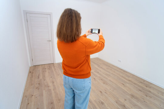 Woman takes photo with smartphone in empty room. Real estate agent documents apartment for sale. House inspection and valuation concept.
