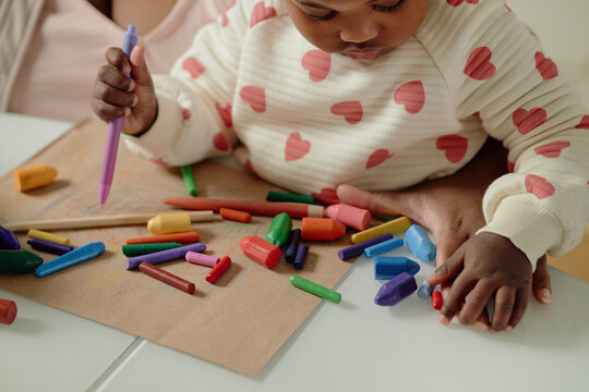Black woman and her toddler drawing with colorful crayons on paper, toddler holding purple pen and touching moms hand