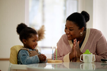 Black woman smiling and clapping hands while sitting with Black toddler in high chair celebrating birthday with small cake and two lit candles on table indoors