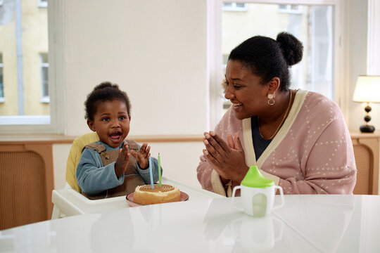 Black woman smiling and clapping hands while sitting beside Black toddler in high chair celebrating birthday with small cake and candle, both showing joyful expressions indoors