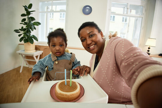 Black woman taking selfie while holding hand of Black toddler sitting in high chair with birthday cake and candles, both looking at camera in bright indoor setting