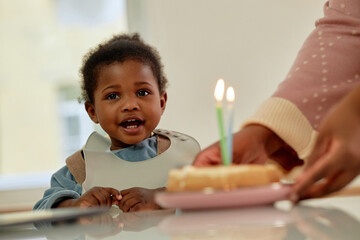Black toddler sitting at table looking toward camera while Black woman presenting birthday cake with lit candles, capturing family celebration and interaction between child and adult