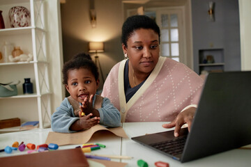 Black woman working on laptop while Black toddler sitting beside her drawing with colored pencils and crayons, both at table in home setting, mother multitasking
