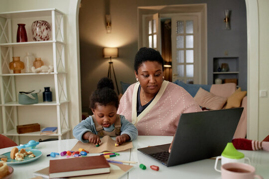 Black woman working on laptop while sitting beside Black toddler drawing with colored crayons at table, both focused on activities in cozy home setting, mother multitasking with child