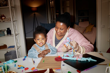 Black woman interacting with Black toddler at table, woman using hand puppet while toddler smiling and looking forward, art supplies and craft materials scattered on tabletop