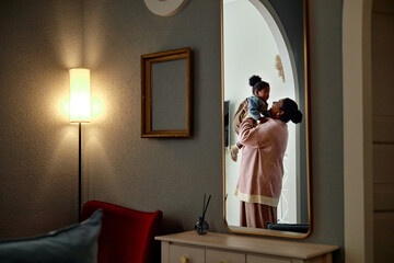 Black woman holding toddler child reflected in mirror, mother lifting young child in cozy home setting, both facing each other, warm family interaction, visible affectionate gesture