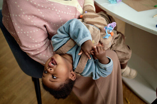 Black woman holding Black toddler upside down on lap, toddler laughing and looking upward, woman sitting at table, both engaging in playful interaction