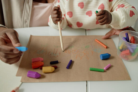 Black woman assisting toddler drawing with colored crayons on brown paper, hands visible holding drawing tools, creative activity involving adult and young child at table