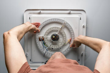 Man installs round LED ceiling light module during home repair, fixing wiring and electronics for safe bright room lighting.