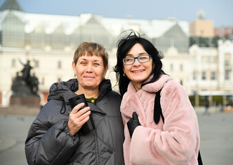 Two Russian friends, 50 and 60 years old, smiling warmly at the camera in the city, sharing joy, confidence, lightness, and the ease of mature female friendship.