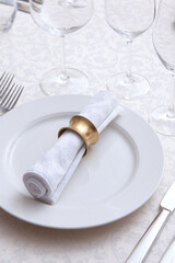 A white napkin with a ring lies on a plate. A napkin ring. Festive table setting. Wine glasses on the table. White plates. VIP lounge. White tablecloth with textured pattern.