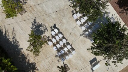 Chairs are neatly arranged in rows under the sun, surrounded by trees and string lights for an upcoming gathering. The space is ready for guests to enjoy.