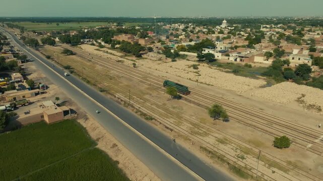 Side-angled aerial clip tracking a train moving parallel to a road with traffic through the Sher Shah village near Multan, Punjab, on a sunny day.