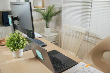 Modern home office workspace presents laptop computer desk for business with monitor, organized papers, green plant, bright blinds, creating comfortable environment for remote work, study or project