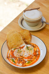 Turkish egg with sourdough bread in a white dish with a cup of hot cappuccino on a wooden table.