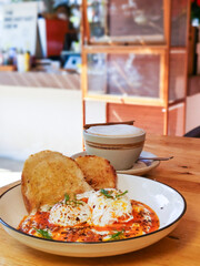 Close up, Turkish egg with sourdough bread in a white dish with a cup of hot cappuccino on a wooden table.