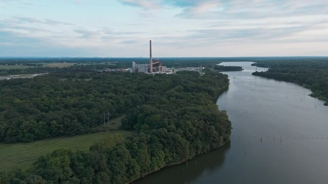 Drone wide overview of Industrial power plant along broad lake mouth curving through wooded hills with smooth water under overcast sky