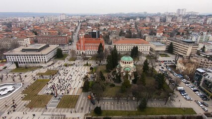 Drone video of Pleven center on March 3, 2019, showing landmarks, the square, the Ossuary, church, municipal building, fountains, and people walking with a long national flag, Pleven, BG,03 March 2019