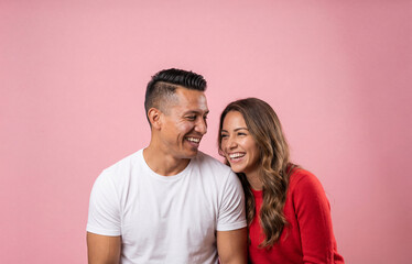 Happy diverse couple laughing together during a fun studio photoshoot. Man and woman in love sharing a candid moment on a pink background.