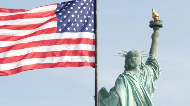 Statue of Liberty on Liberty Island, New York, United States. Cooper monument from behind, democracy and patriotism symbol near Manhattan, NYC USA. American flag waving in wind. Independence concept.