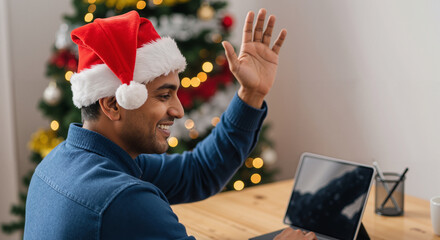 Smiling man in Santa hat waves during a festive Christmas video call on a tablet, celebrating holidays virtually.