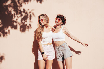 Two young beautiful smiling hipster female in trendy summer white t-shirt and shorts clothes. Sexy carefree women posing in the street. Positive models having fun, hugging and going crazy, near wall