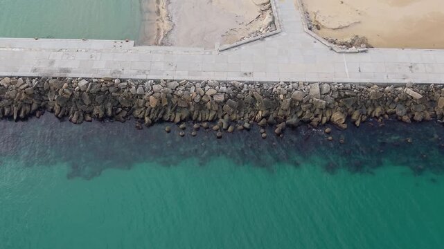 Drone footage captures a concrete breakwater surrounded by dark tetrapods in turquoise coastal water, showing calm waves, shoreline details and peaceful marine atmosphere.