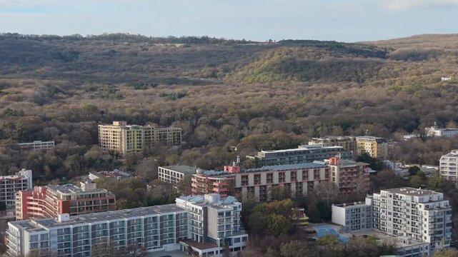 Aerial drone footage showing residential buildings surrounded by wooded hills and valleys, highlighting urban development integrated with natural landscape under soft daylight.