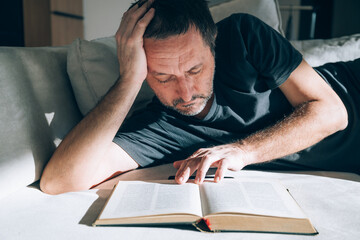 Authentic middle-aged man lying on couch, looking exhausted and deep in thought while reading an open book, hand on forehead. Perfect for overthinking, mental fatigue and literature.