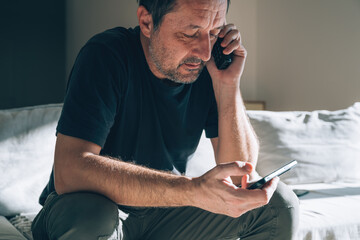 Authentic middle-aged caucasian man looking seriously worried while talking on landline phone and checking smartphone. Stress, bad news, crisis, debt, mental health themes.
