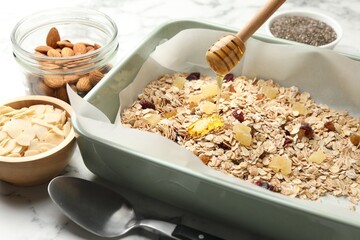 Making granola. Pouring honey onto oat flakes and dried fruits at white marble table, closeup