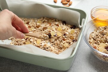 Making granola. Woman adding oat flakes with dried fruits into baking dish at grey textured table, closeup