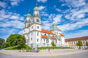 Naklejka premium St. Lorenz Basilica stands majestically in Kempten, Bavaria, showcasing baroque architecture. Visitors admire the structure against a vibrant sky, surrounded by greenery and historical buildings.