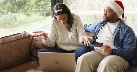 Diverse couple reacting after completing card payment on laptop while wearing Santa hat on couch