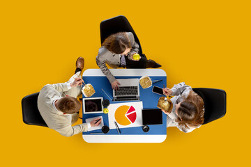 Business team eating noodles and reviewing charts around laptop at blue desk in overhead angle. Concept of food delivery apps, business workflow, education materials and startup teamwork.