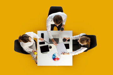 Team reviewing charts and devices during coffee break at white desk in overhead angle. Concept of business workflow, marketing materials, education content and office routines.