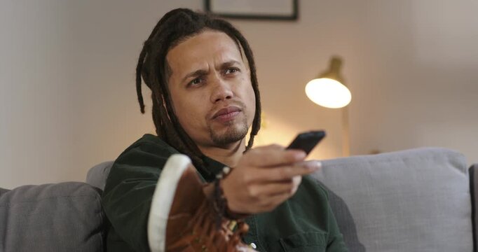 Middle-aged African American man pressing remote control on sofa in living room scanning channels