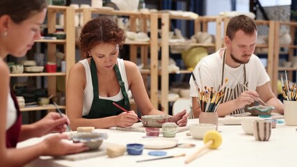 Three craftsperson are busy painting clay products. They are sitting near desktop, using brush and paints, painting product made of raw clay. Working together in pottery workshop. 