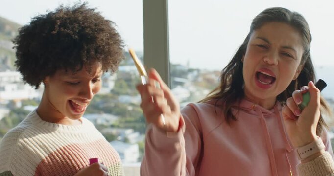 Diverse female friends grabbing nail polish bottle and makeup brush lip-syncing for fun at home