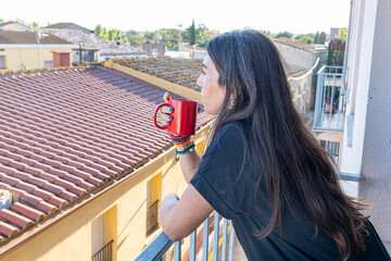 Woman standing on an apartment balcony drinking from a red mug, looking over city rooftops