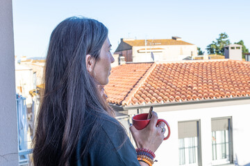 Woman enjoying a peaceful morning, drinking from a red mug while observing the city view from her home